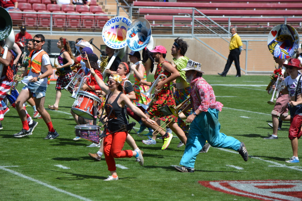 Eine Gruppe von Menschen im Freien mit Trommeln, verschiedenen Instrumenten, Stühlen, Treppen und einem Zaun im Hintergrund.