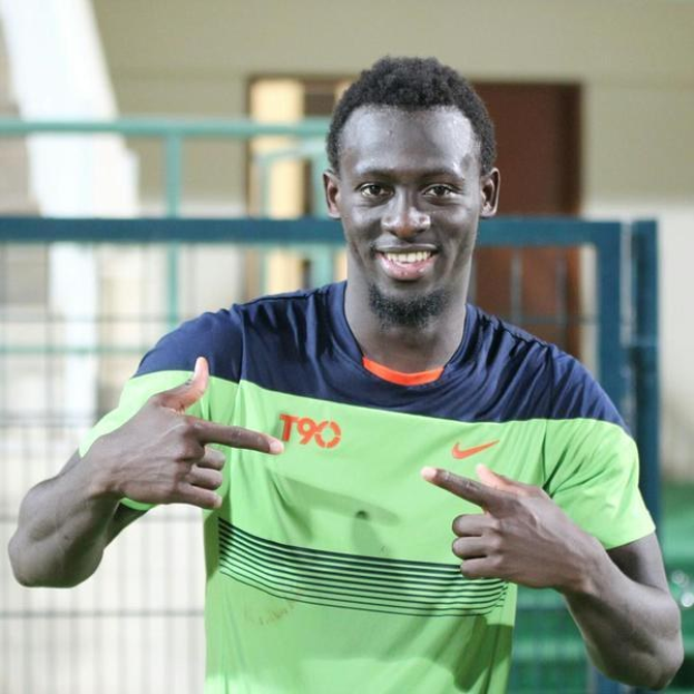 Ein Mann mit einem grünen und blauen Hemd lächelt und zeigt auf die Kamera, mit einem unscharfen Geländer, einer Wand und einer Tür im Hintergrund.