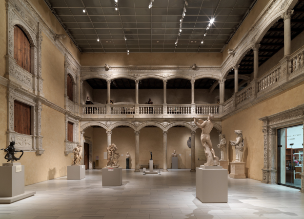 Ein geräumiger Museumsgalerie mit zahlreichen Statuen und Skulpturen auf Sockeln, mit architektonischen Elementen wie Wänden, Säulen, Geländern und einer beleuchteten Decke und einigen Besuchern, die die Kunstwerke betrachten.