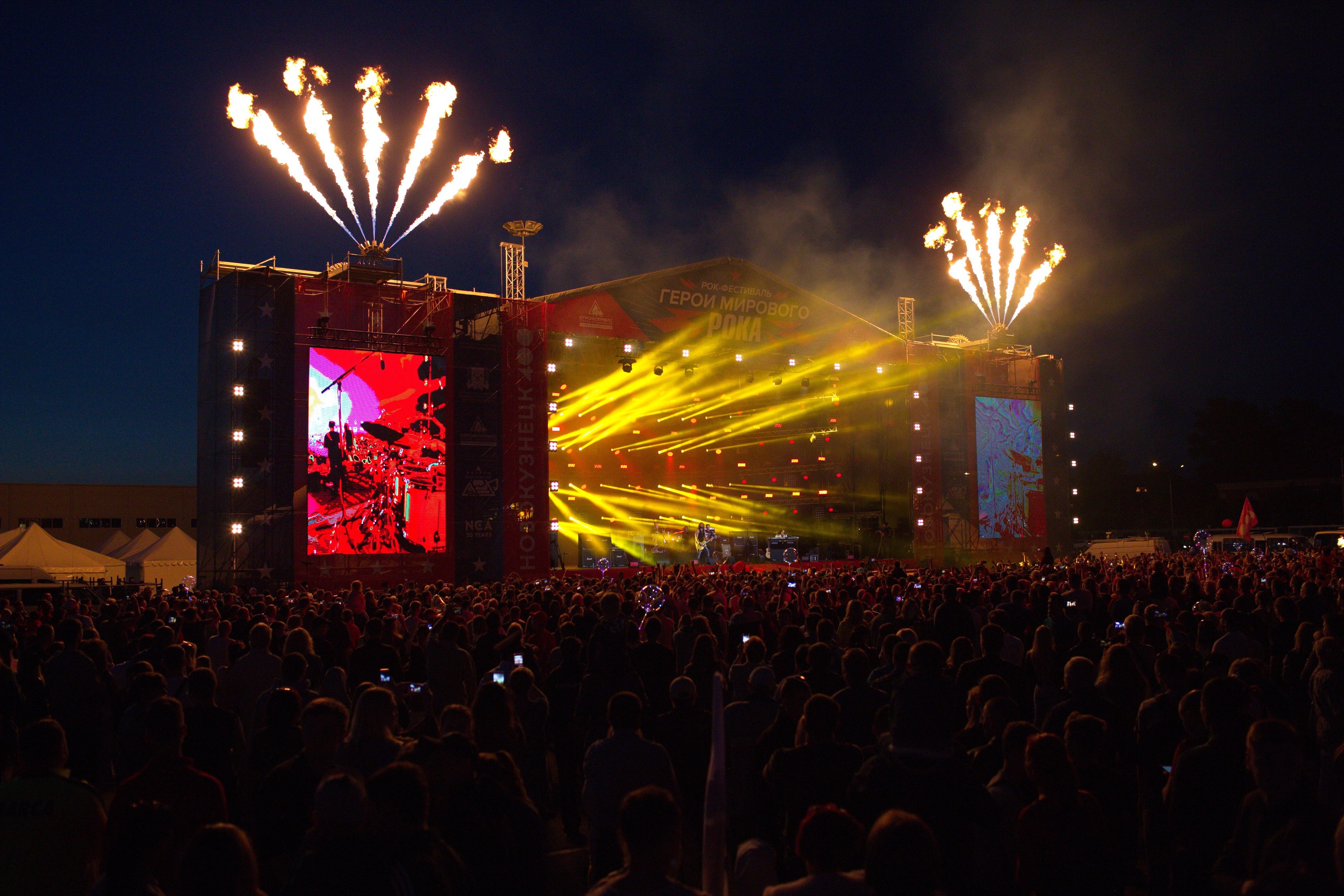Eine große Menge bei einem Musikfestival schaut Performern auf einer hell erleuchteten Bühne zu, mit Zelten links und Feuerwerk am dunklen Himmel dahinter.