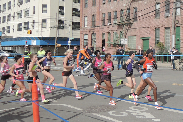 Gruppe von Läufern bei einem Marathon auf einer Straße mit Gebäuden, Strommasten, Verkehrszeichen, Schildern, Fußgängern, einem Radfahrer, Bäumen und einem klaren blauen Himmel.