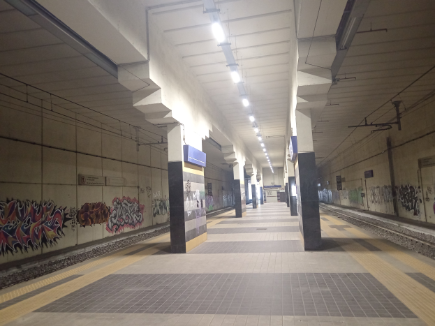 Eine U-Bahn-Station mit Graffiti an den Wänden, beleuchtet von Deckenlampen, mit Säulen, Tafeln und einer Eisenbahnschiene auf der rechten Seite.