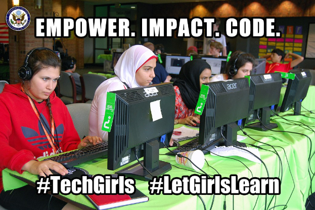 Gruppe junger Frauen an einem Tisch mit Computern, Tastaturen und Bühchern, die Kopfhörer tragen und von Stühlen umgeben sind, mit einem Logo und Text an der Wand, der "empower, impact, code, techgirls" lautet.