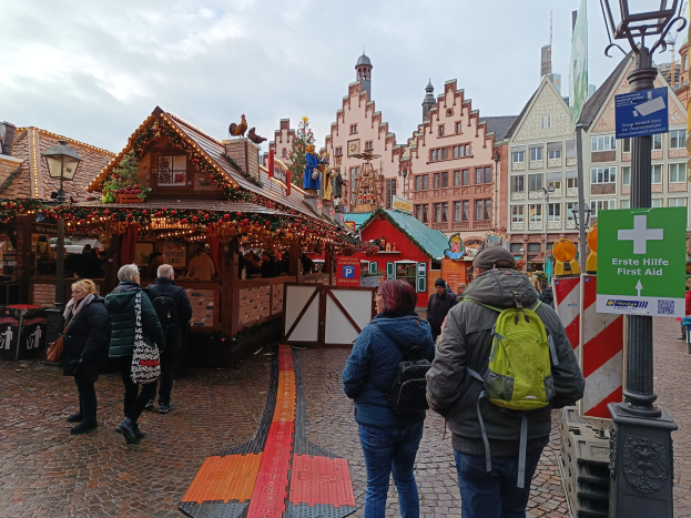 Eine Gruppe von Menschen geht eine Kopfsteinpflasterstraße entlang neben einem Weihnachtsmarkt in Nürnberg, Deutschland, mit Laternenmasten, Texttafeln und Gebäuden im Hintergrund unter einem bewölkten Himmel.