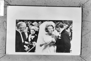 Ein Schwarz-Weiß-Foto von einem Brautpaar, das sich die Ringe austauscht, umgeben von einer Gruppe von Menschen im Hintergrund, wobei der Mann einen Anzug und die Frau ein Kleid trägt.