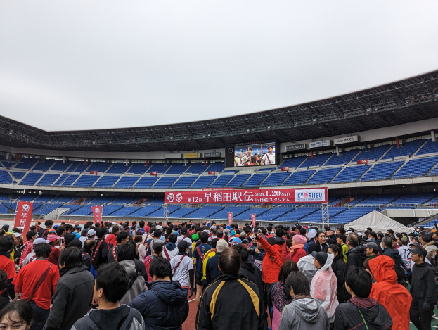 Große Menschenmenge vor einem Stadion bei einem Event mit Bannern, Stühlen und einem Bildschirm im Hintergrund während der Eröffnungsfeier der Olympischen Spiele Tokyo 2020