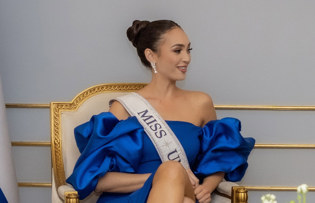 Frau in einem blauen Kleid mit einem "Miss Universe 2019" -Band, die lächelt, während sie auf einem Stuhl sitzt, umgeben von Blumen und einer Wand.