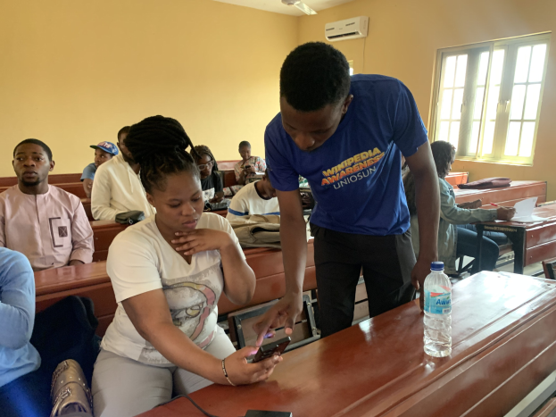 Eine Gruppe von Schülern sitzt an Tischen in einem Klassenzimmer, während ein Mann mit einem Mobiltelefon in der Hand steht; Flaschen und andere Gegenstände befinden sich auf den Tischen, und ein Klimagerät und ein Fenster sind im Hintergrund zu sehen.