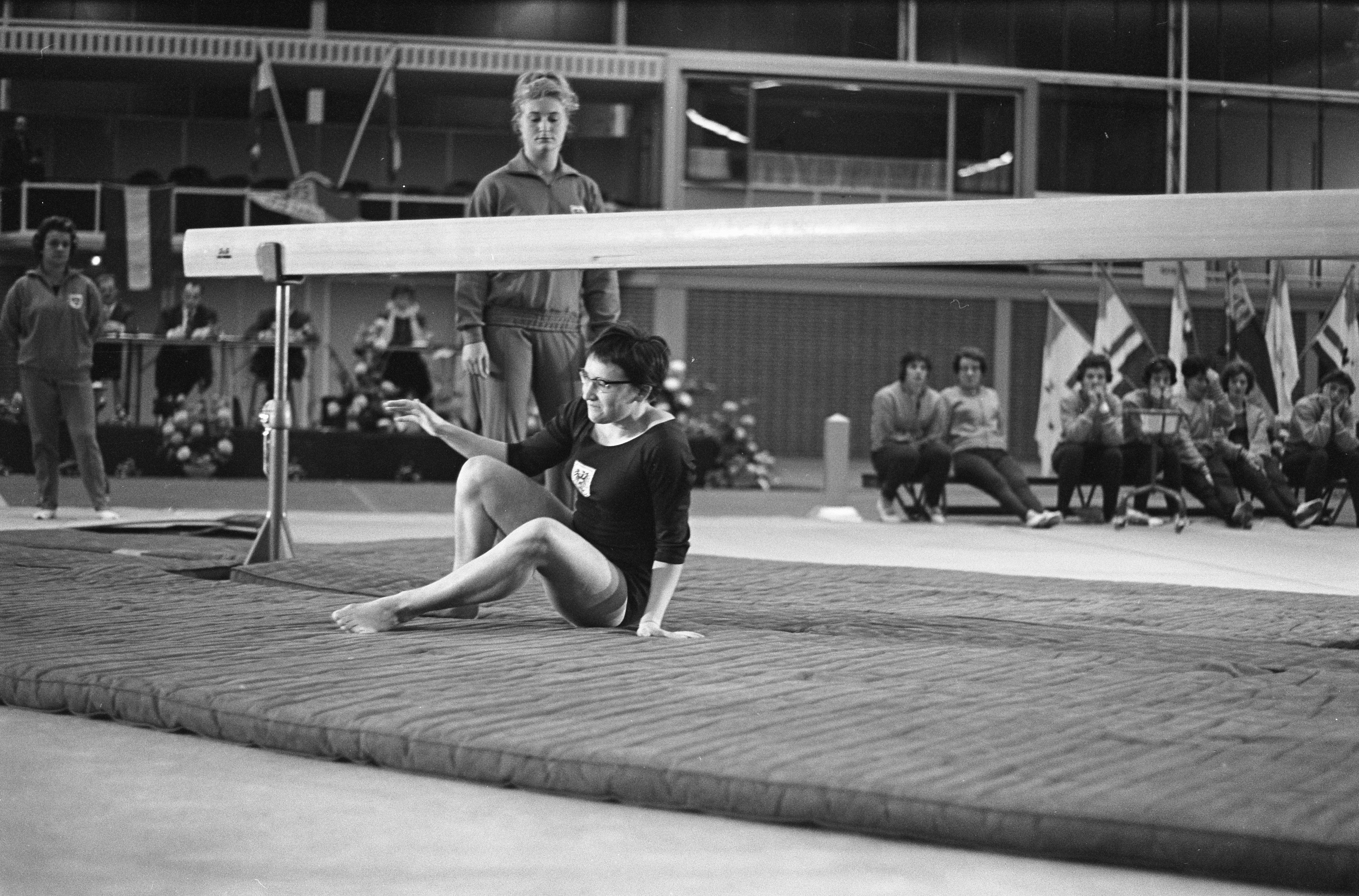 Eine Frau in einem schwarzen und weißen Turnoutfit führt eine Übung auf einem Balancebalken in einer Turnhalle durch, mit Zuschauern und Fahnen im Hintergrund.