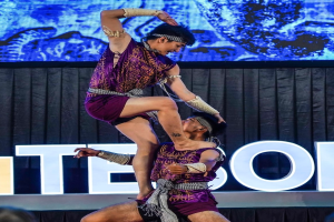 Ein Mann und eine Frau in lila Kleidern führen Akrobatik auf der Bühne vor einem Publikum auf, wobei der Mann die Frau in seinen Armen hält.