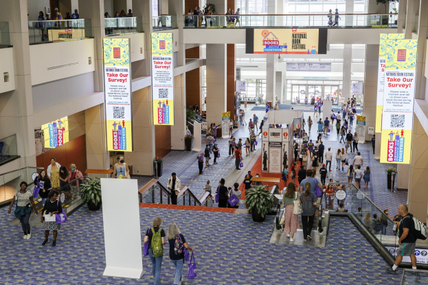 Eine große Menschenmenge, die durch ein großes Gebäude mit Bannern, Topfpflanzen, Rolltreppen, Geländern, Säulen, bedruckten Schildern, Glastüren und anderen Gegenständen geht, wobei einige Taschen tragen, wahrscheinlich an der Nationalen Buchmesse teilnehmen.