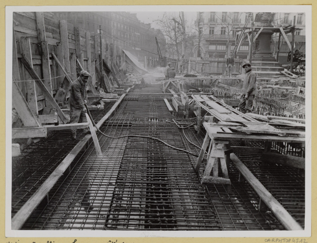 Schwarzes und weißes Foto von Männern bei der Arbeit auf einer Baustelle mit Gebäuden, Bäumen, Masten, Drähten im Hintergrund und Text unten.