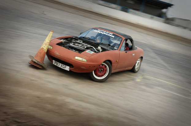 Roter Wagen auf einer Rennstrecke mit einem Verkehrskegel vorne, unscharfer Hintergrund, der hohe Geschwindigkeit andeutet.