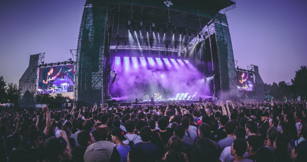 Eine große Menschenmenge auf einem Musikfestival schaut auf einer hell erleuchteten Bühne mit Instrumenten und Bildschirmen, die vor einem Gebäude, Bäumen und einem klaren blauen Himmel stehen.