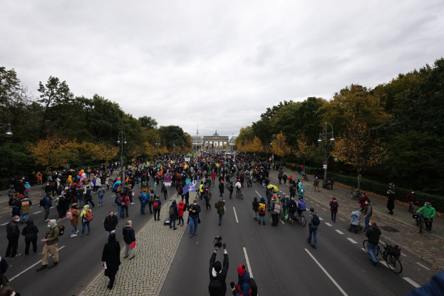 Eine große Gruppe von Menschen marschiert auf einer baumbestandenen Straße in Berlin, mit Kameras in der Hand, vor einem Gebäude und einem klaren Himmel