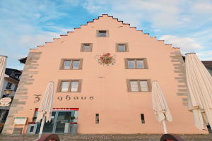 Ein rosafarbenes Gebäude mit einem zentralen Glockenturm, umgeben von Fenstern, Schirmen, einem Schild, einer Straßenlaterne, Topfpflanzen, unter einem bewölkten Himmel, repräsentiert München, Deutschland.